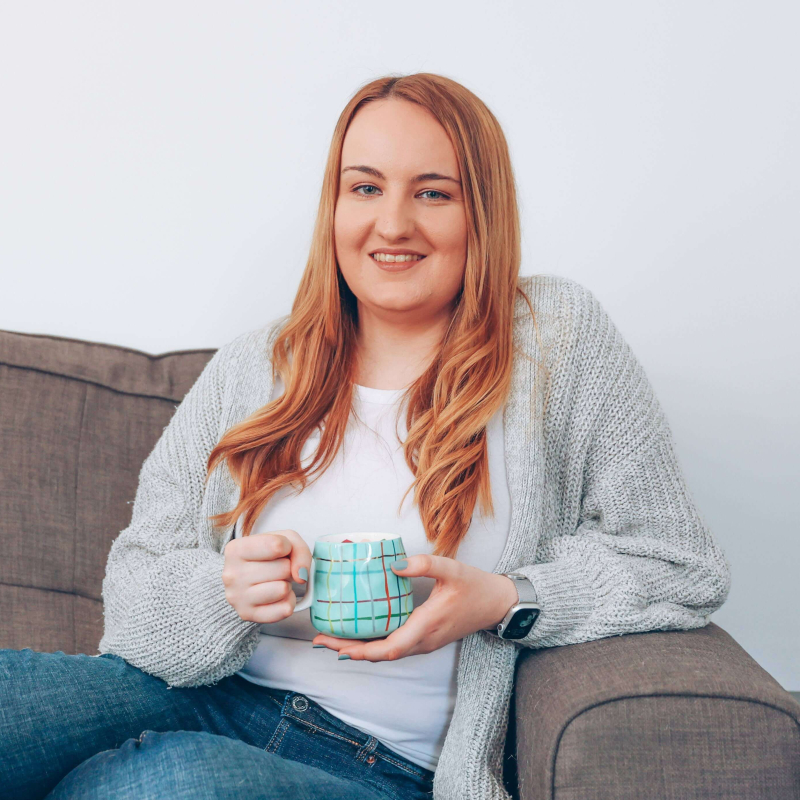 A photo of Holly Pryce; a woman with long blonde hair. She is sitting on a grey sofa holding a cup of tea.