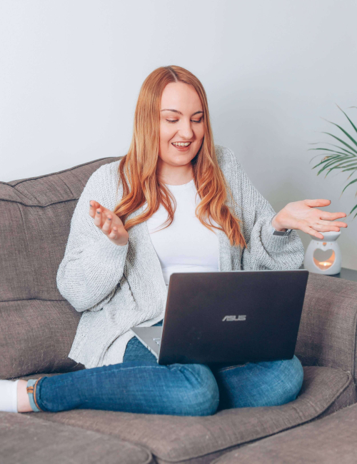 A photo of Holly Pryce; a woman with long blonde hair. She is sitting with a laptop on her lap, gesturing with her hands.