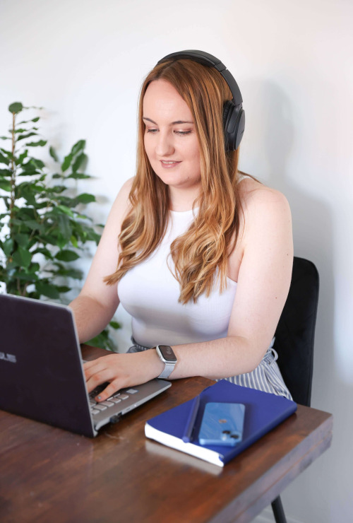 A photo of Holly Pryce; a white woman with blonde hair. She is sitting at a table working on a laptop, wearing over ear headphones.