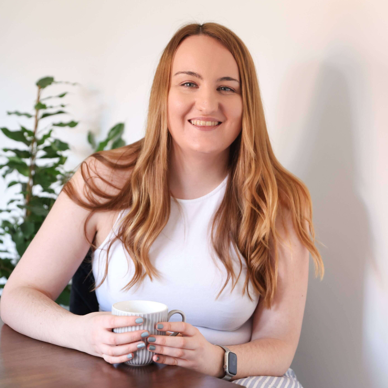 A photo of Holly Pryce; a white woman with blonde hair. She is looking at the camera while smiling and holding a mug.